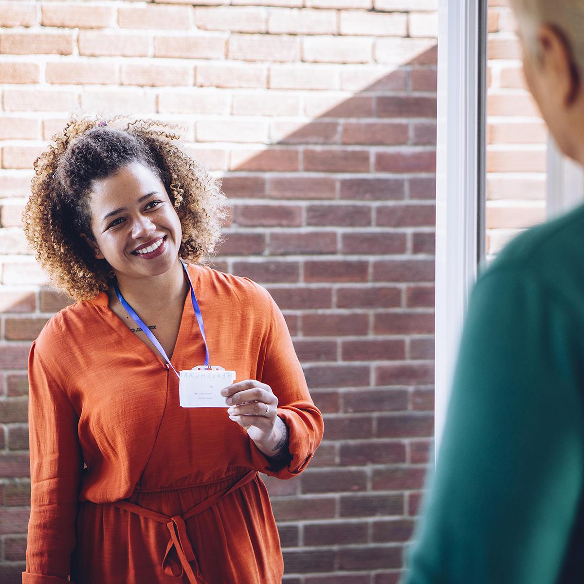 Photo of a smiling young Black woman in an orange dress, holding her social worker ID out to greet an elderly man